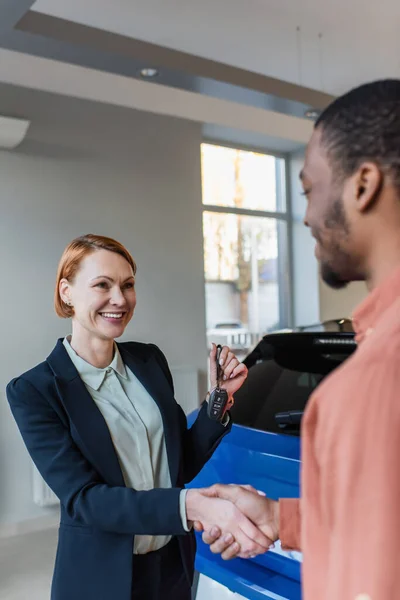 Feliz concesionario de coches sosteniendo la llave y estrechando la mano con borrosa cliente afroamericano - foto de stock