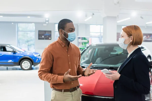 Hombre afroamericano en máscara médica hablando con gerente de sala de exposición de coches - foto de stock