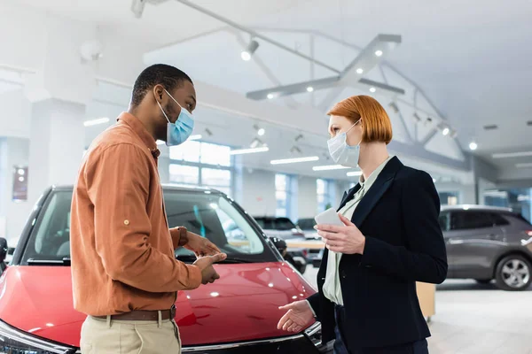 African american man in medical mask pointing with hands at car near car dealer — Stock Photo