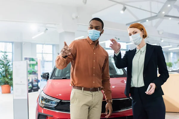 Concessionnaire automobile et client afro-américain dans les masques médicaux pointant avec les doigts dans la salle d'exposition de voiture — Photo de stock