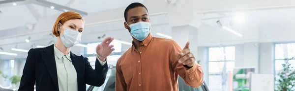 African american man and car dealer in medical masks pointing with fingers in car showroom, banner — Stockfoto