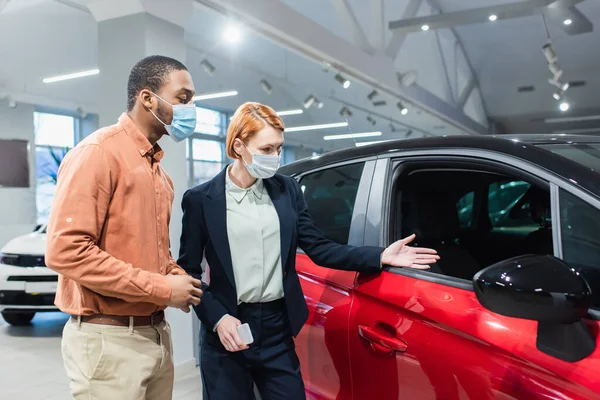 Car dealer in medical mask pointing with hand at auto near african american man — Stock Photo