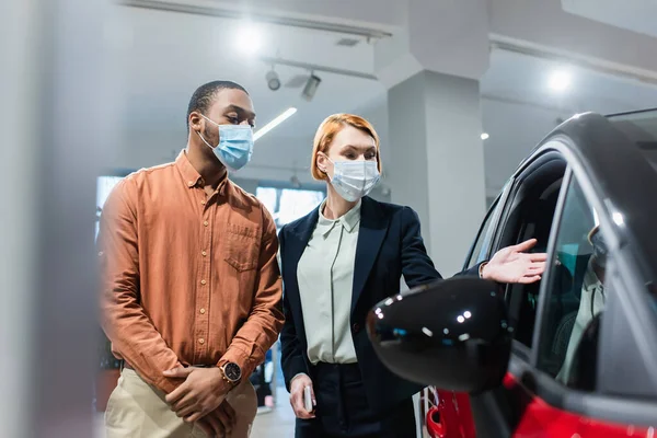 Concessionário de automóveis em máscara médica apontando para o carro perto de cliente afro-americano — Fotografia de Stock
