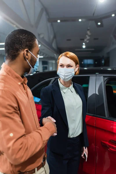 Concesionario de coches y cliente afroamericano en máscaras médicas estrechando las manos cerca de coche - foto de stock