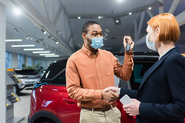 African american man with key and car dealer in medical masks shaking hands in car showroom — Stock Photo