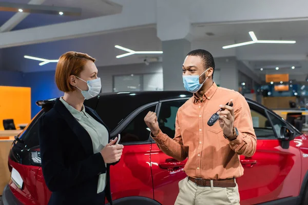 Hombre afroamericano emocionado en máscara médica sosteniendo la llave y mostrando el gesto de ganar cerca de coche y vendedor - foto de stock