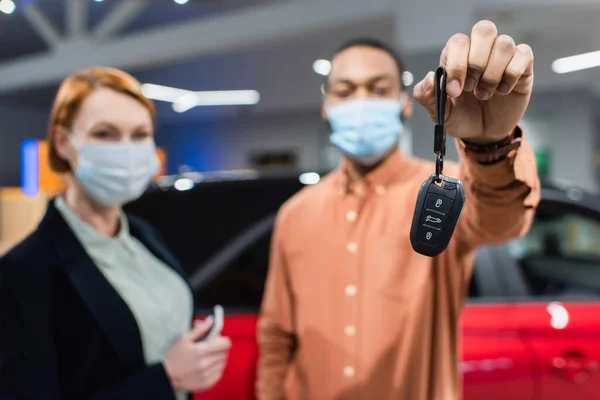 Selective focus of key in hand of blurred african american man in medical mask standing near car dealer — Fotografia de Stock