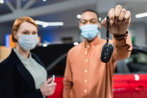 Selective focus of key in hand of blurred african american man in medical mask standing near car dealer — Stock Photo