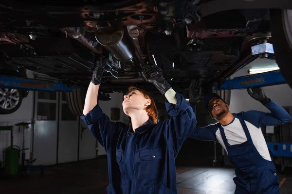 Interracial technicians inspecting bottom of lifted auto in car service — Stockfoto