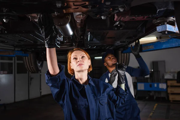 Multiethnic mechanics checking bottom of lifted car in workshop — стоковое фото