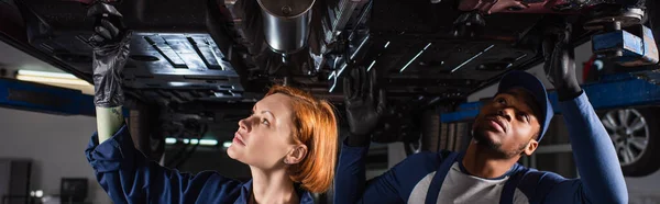 Interrassische Mechaniker, die Diagnose des angehobenen Fahrzeugs im Autoservice machen, Banner — Stockfoto