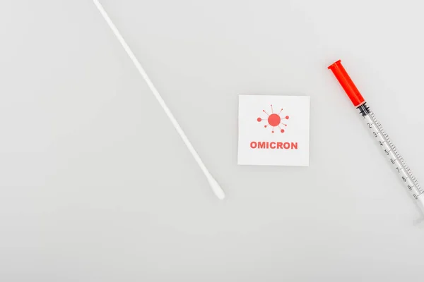 Top view of card with red omicron lettering and bacteria icon near syringe and swab test on grey background — Foto stock