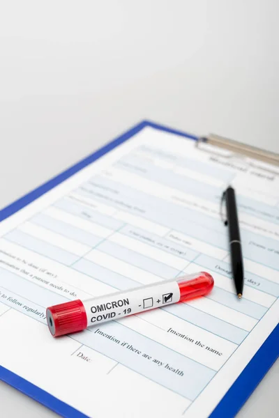 Test tube with covid-19 omicron variant positive result near clipboard and pen on grey background — стоковое фото