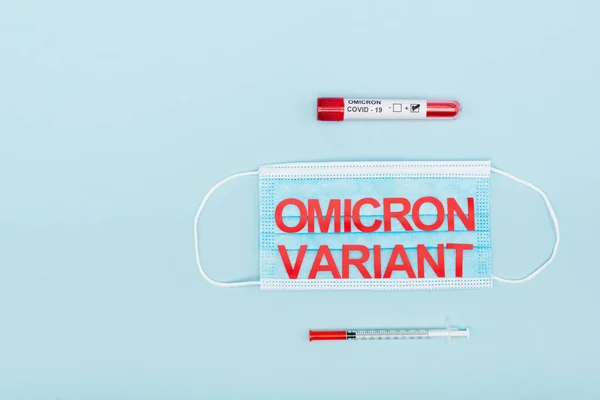 Top view of medical mask with omicron variant lettering near test tube and syringe on blue — Fotografia de Stock