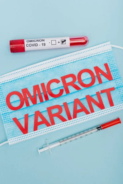 Top view of medical mask with red omicron variant lettering near syringe and test tube with positive result on blue — Foto stock