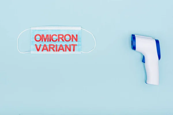 Top view of medical mask with red omicron variant lettering near pyrometer on blue — Stock Photo