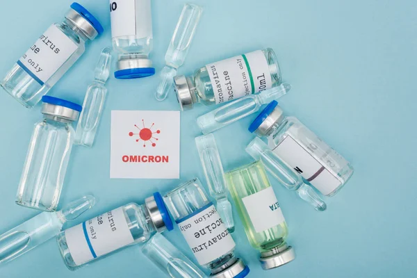 Top view of white card with red omicron lettering and bacteria near vaccine vials and ampoules on blue — стоковое фото