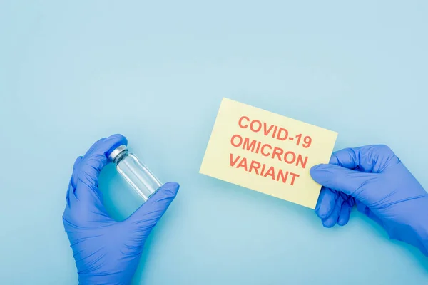 Partial view of doctor in latex gloves holding vaccine vial and card with covid-19 omicron variant lettering on blue — Foto stock