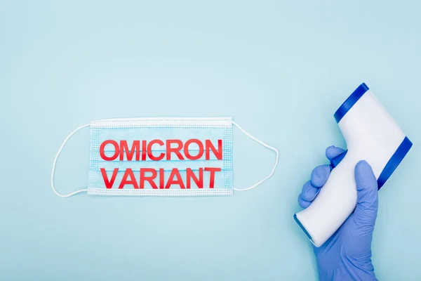 Cropped view of doctor in latex glove holding pyrometer near medical mask with omicron variant lettering on blue — Photo de stock