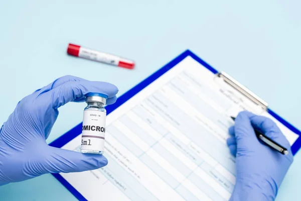 Cropped view of immunologist in latex gloves holding covid-19 omicron variant vaccine and writing on blurred clipboard on blue — Fotografia de Stock