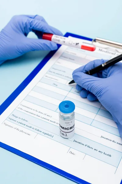 Cropped view of doctor in latex gloves writing on clipboard near vial with covid-19 omicron variant vaccine on blue — Foto stock