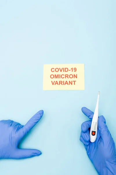 Partial view of doctor with electronic thermometer pointing at card with covid-19 omicron variant lettering on blue — Stockfoto