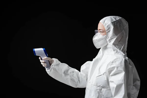 Vue latérale du médecin en combinaison Hazmat, lunettes et masque médical tenant pyromètre isolé sur noir — Photo de stock