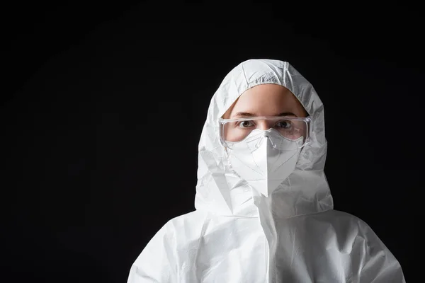 Doctor in goggles, medical mask and white hazmat suit looking at camera on black, omicron variant concept — стоковое фото