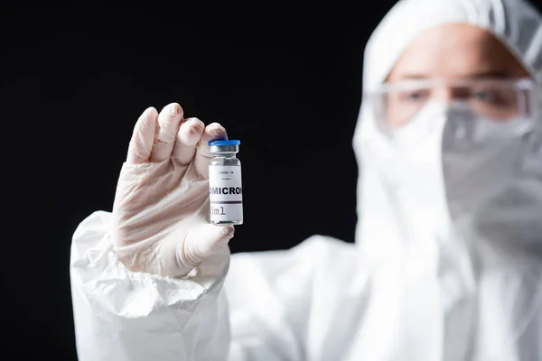 Blurred immunologist in white hazmat suit holding covid-19 omicron variant vaccine isolated on black — Stock Photo