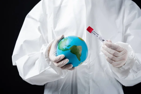 Cropped view of scientist in white hazmat suit holding globe and covid-19 omicron variant test isolated on black — Stock Photo