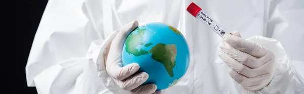 Cropped view of doctor in hazmat suit holding globe and covid-19 test tube isolated on black, banner — Stockfoto