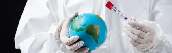 Cropped view of doctor in hazmat suit holding globe and covid-19 test tube isolated on black, banner — стоковое фото
