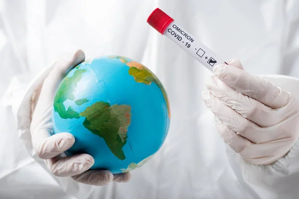 Partial view of scientist in hazmat suit and latex gloves holding globe and positive covid-19 omicron variant test — Stockfoto