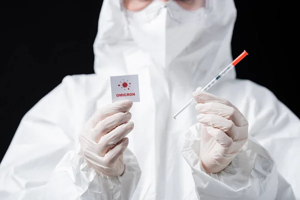Partial view of doctor in hazmat suit holding syringe and card with omicron lettering and bacteria icon isolated on black — Stockfoto
