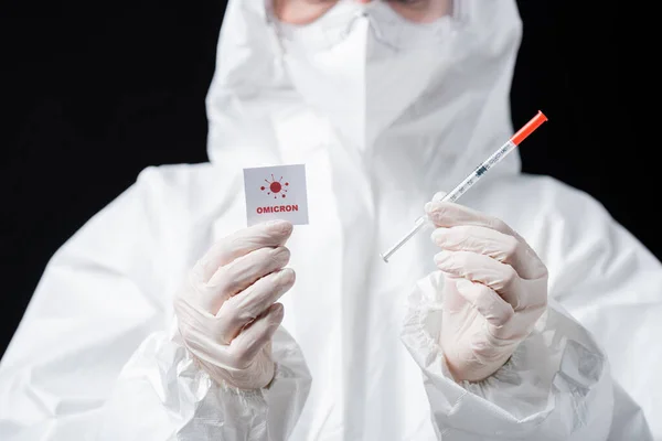 Partial view of doctor in hazmat suit holding syringe and card with omicron lettering and bacteria icon isolated on black — стоковое фото