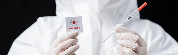 Partial view of scientist in latex gloves holding syringe and card with omicron lettering isolated on black, banner — Stock Photo