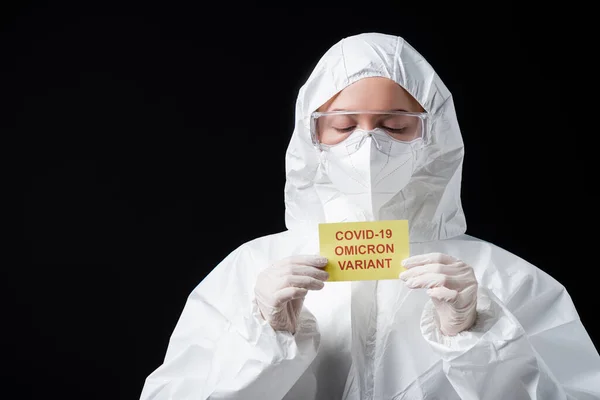 Scientist in white hazmat suit and goggles holding card with covid-19 omicron variant lettering isolated on black — Photo de stock