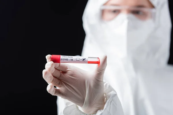 Selective focus of positive covid-19 omicron variant test in hand of blurred microbiologist in hazmat suit isolated on black — Stock Photo