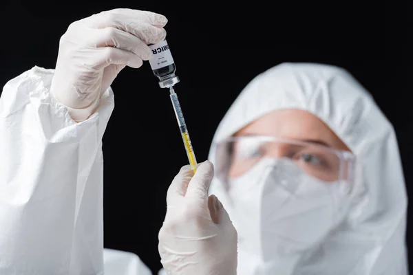 Blurred immunologist in hazmat suit holding syringe and covid-19 omicron variant vaccine isolated on black — Foto stock