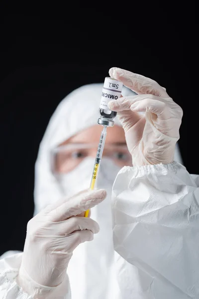 Blurred immunologist in hazmat suit holding syringe and omicron variant vaccine isolated on black — Stock Photo