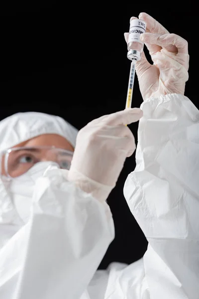 Blurred doctor in personal protective equipment holding syringe and omicron variant vaccine isolated on black — стоковое фото