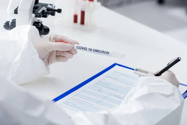 Partial view of blurred doctor holding covid-19 omicron variant test tube while writing on clipboard in lab — Stock Photo