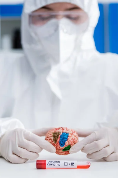 Blurred doctor in hazmat suit holding brain model near positive covid-19 omicron variant test — Stock Photo