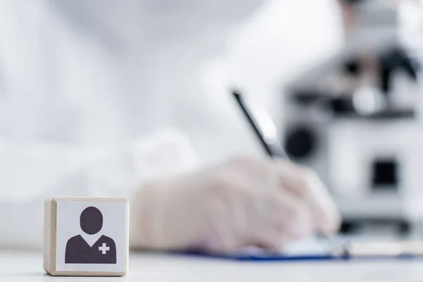 Selective focus of cube with medical icon near scientist writing near microscope on blurred background, omicron variant concept — Stock Photo