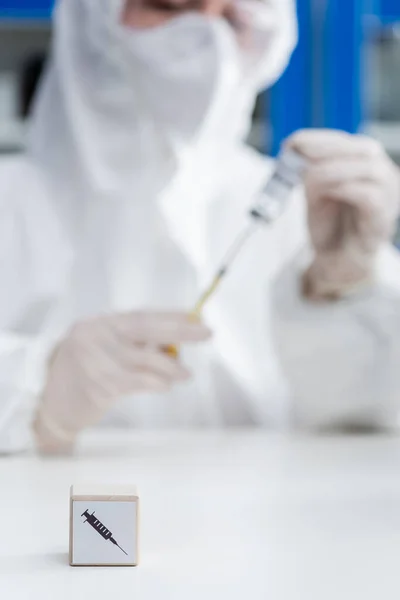 Cropped view of blurred immunologist in hazmat suit holding vaccine near cube with syringe icon, omicron variant concept — Foto stock