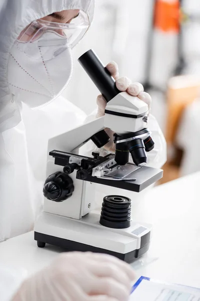 Biólogo en traje de materiales peligrosos, gafas y máscara médica trabajando con microscopio en laboratorio, concepto de variante omicrónica - foto de stock