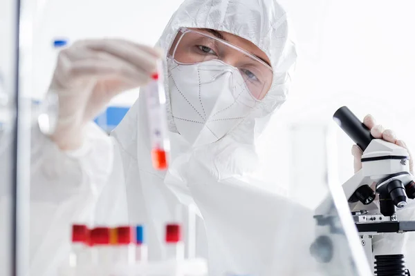 Scientist in goggles, medical mask and hazmat suit holding blurred test tube near microscope in lab - foto de stock