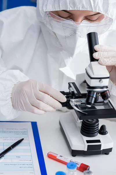 Científico en traje de materiales peligrosos que trabaja con microscopio cerca de tubo de ensayo y portapapeles en laboratorio - foto de stock