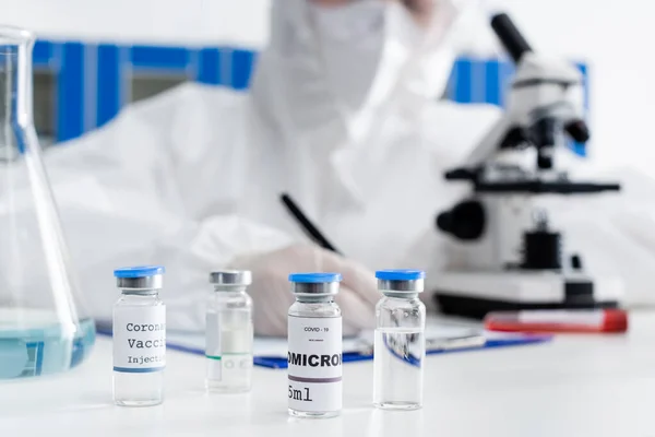 Partial view of blurred doctor writing on clipboard near microscope and vials with covid-19 omicron vaccine — Foto stock