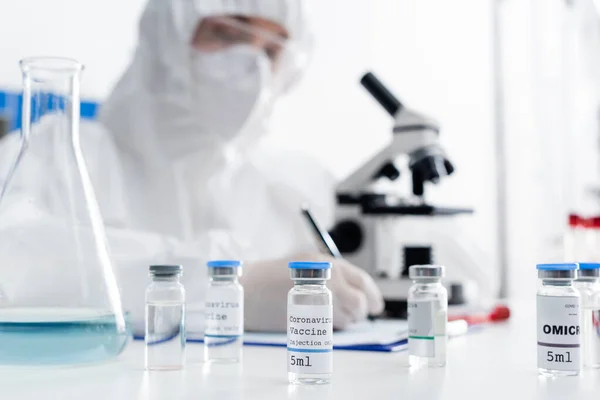 Selective focus of vials with coronavirus vaccine near scientist writing near microscope on blurred background — стоковое фото